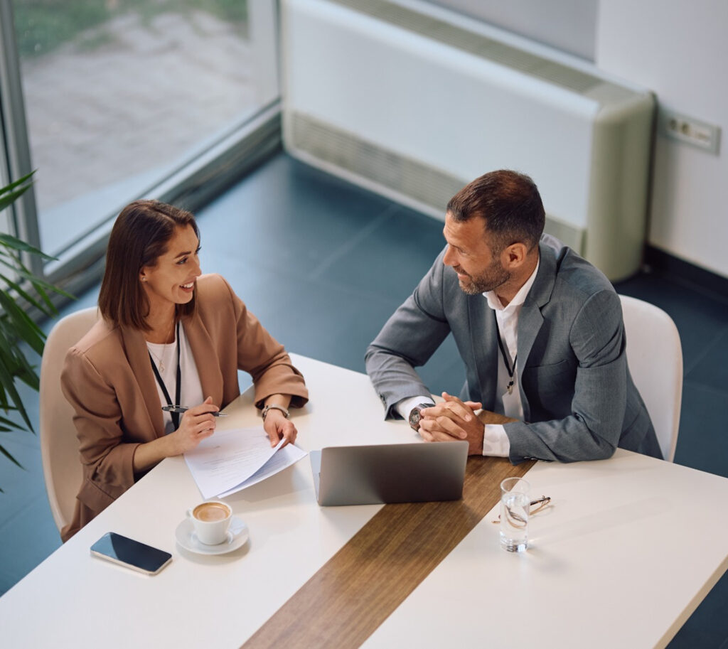 Profissionais em ambiente corporativo: homem de terno cinza e mulher de blazer bege conversam, mesa com documentos, laptop e café; presença do IDOR e Faculdade IDOR